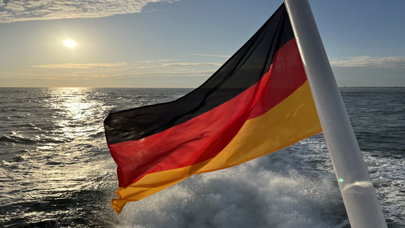 Am Heck eines fahrenden Schiffes weht die Flagge Deutschlands. Im Hintergrund ist eine niedrig stehende Sonne über dem Horizont zu sehen. Das Sonnenlicht spiegelt sich auf der Wasseroberfläche. Am Heck eines fahrenden Schiffes weht die Flagge Deutschlands. Im Hintergrund ist eine niedrig stehende Sonne über dem Horizont zu sehen. Das Sonnenlicht spiegelt sich auf der Wasseroberfläche.