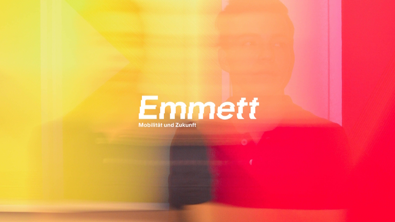 Schriftgrafik mit dem Text „Emmett. Mobilität und Zukunft.“ Im Hintergrund sind ein gelb-roter Farbverlauf sowie die Spiegelung einer Person zu erkennen. Schriftgrafik mit dem Text „Emmett. Mobilität und Zukunft.“ Im Hintergrund sind ein gelb-roter Farbverlauf sowie die Spiegelung einer Person zu erkennen.