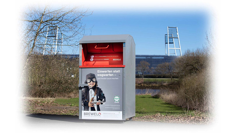 Altkleidercontainer mit Abdruck Einwerfen statt wegwerfen. im Hintergrund ein Fußballstadion. Altkleidercontainer mit Abdruck Einwerfen statt wegwerfen. im Hintergrund ein Fußballstadion.