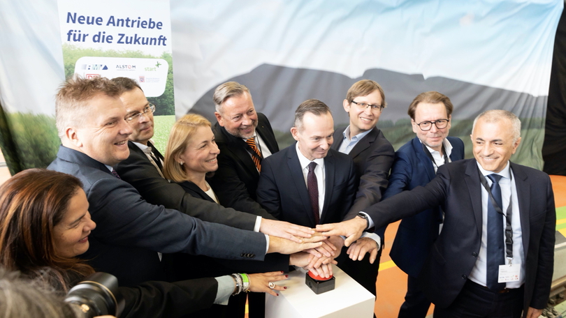 Mehrere Personen drücken symbolisch gemeinsam auf einen großen roten Knopf und lächeln. Mehrere Personen drücken symbolisch gemeinsam auf einen großen roten Knopf und lächeln.