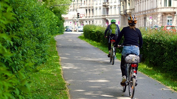 Zum Artikel Zwei Radfahrer auf einem städtischen Fahrradweg umgeben von Bäumen und Sträuchern
