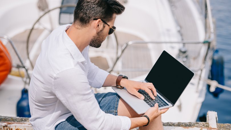 Junger Mann mit Sonnenbrille der mit einem Laptop auf einem Boot sitzt Junger Mann mit Sonnenbrille der mit einem Laptop auf einem Boot sitzt