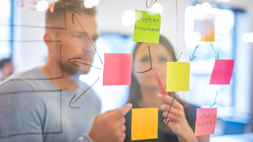 Zwei Personen arbeiten gemeinsam an einer Mindmap aus verschiedenfarbigen Klebezetteln, welche an einer transparenten Tafel angebracht sind. Zwei Personen arbeiten gemeinsam an einer Mindmap aus verschiedenfarbigen Klebezetteln, welche an einer transparenten Tafel angebracht sind.